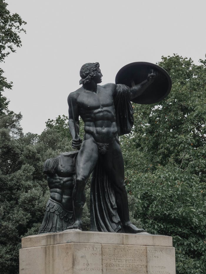 A striking bronze statue of a hero holding a shield, set against a backdrop of green trees.
